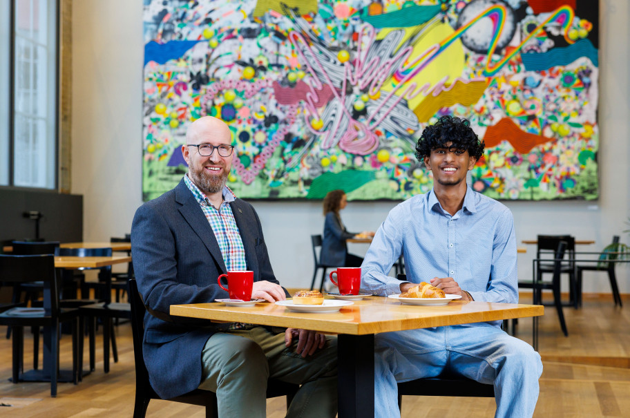 Zwei Männer sitzen vor einem bunten Gemälde an einem Tisch mit Kaffee