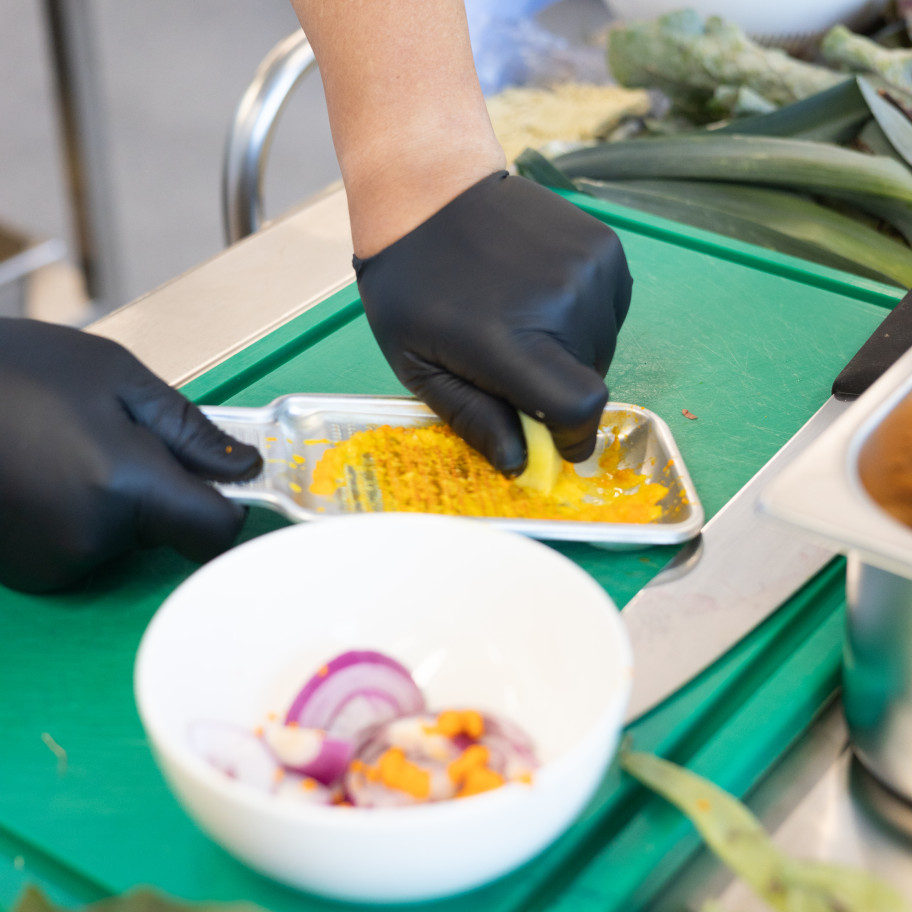 Hand mit Gastrohandschuh reibt Ingwer auf einer kleinen Reibe