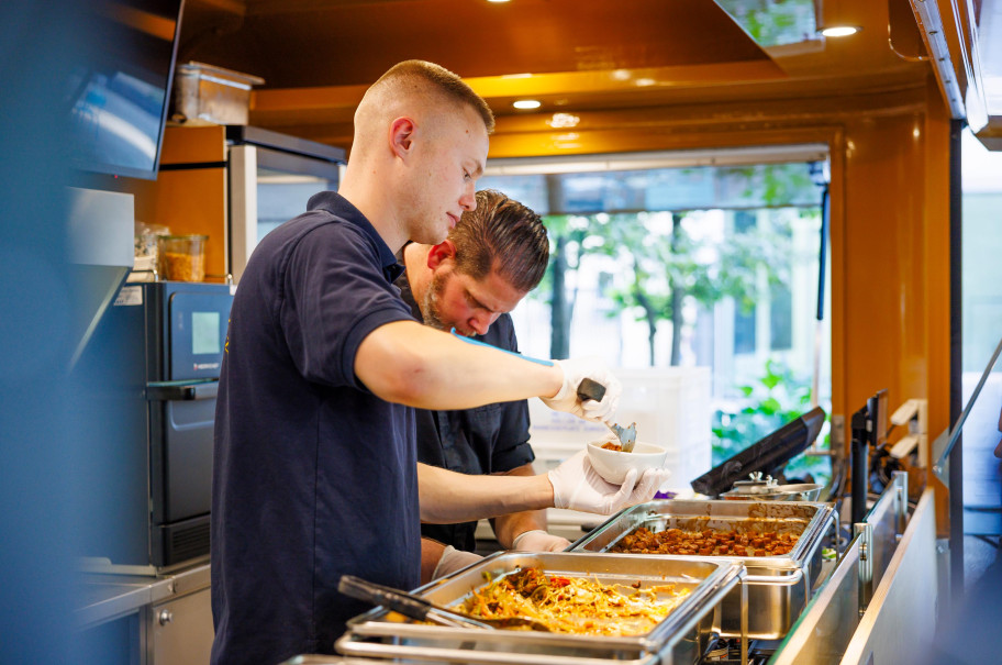 zwei Männer in einem Foodtruck mit blauen Shirts bereiten Essen zu