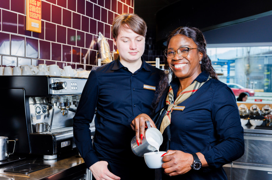 Frau lächelt in die Kamera und zeigt einem Auszubildenden Systemgastronomen Latteart