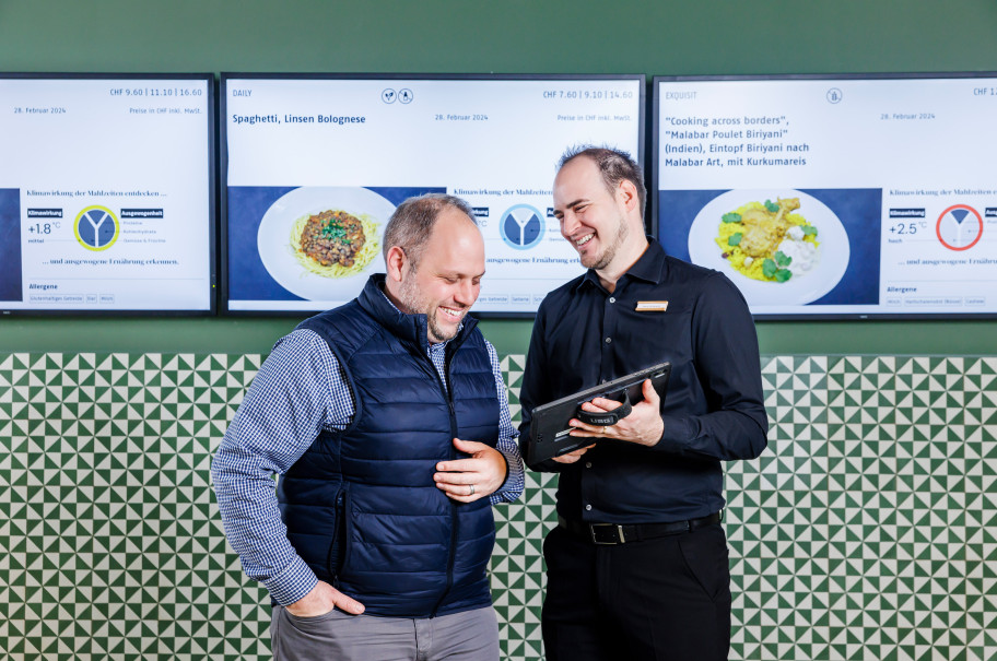 Zwei Männer unterhalten sich vor Screens, die Klimawirkung von Gerichten aufzeigen