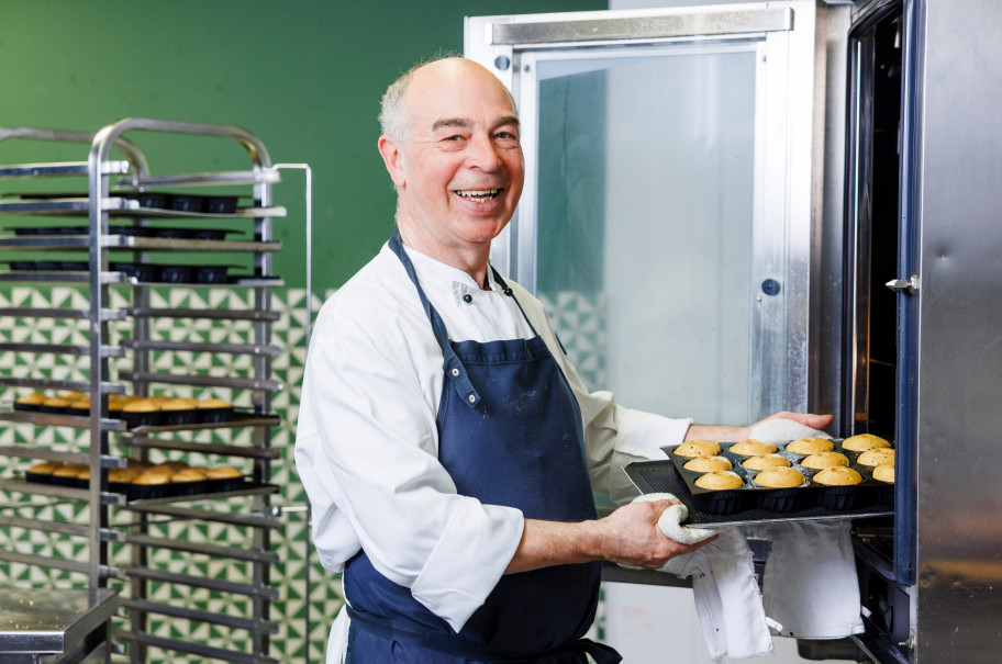 Konditor mit Schürze zieht lachend ein Blech hausgemachte Muffins aus dem Ofen