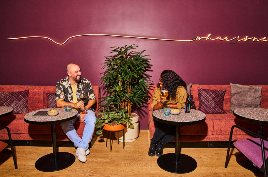 Zwei Personen sitzen an Tischen vor einer bunten Wand und lachen sich an