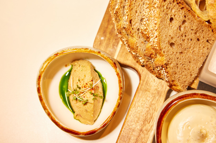 Schüssel mit cremigem Aufstrich, garniert mit Kresse und grüner Sauce, daneben mehrere Scheiben rustikales Brot auf einem Holzbrett