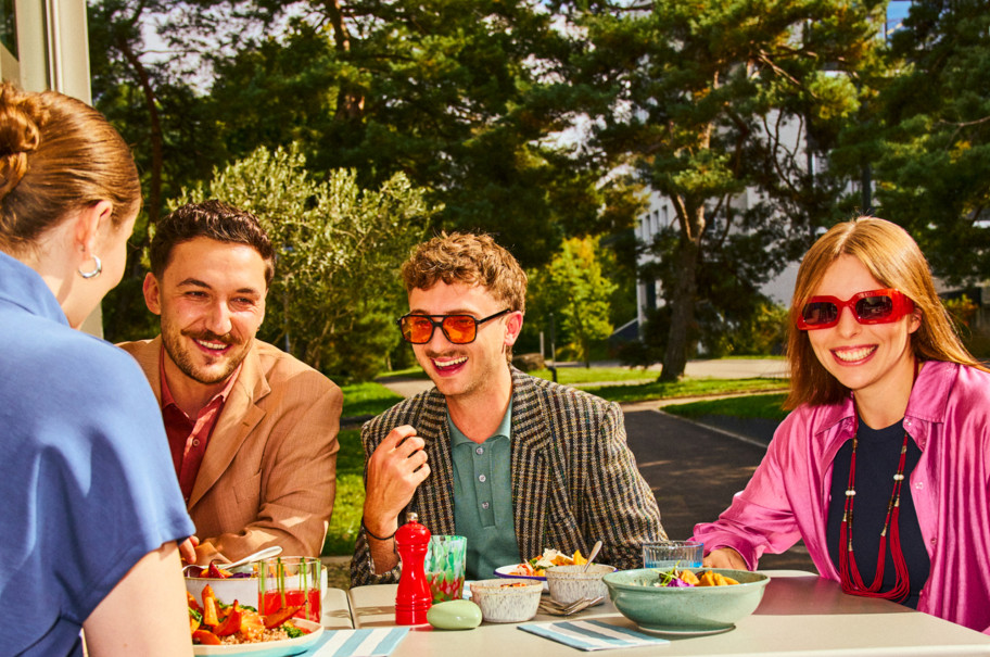 Vier Personen sitzen an einem gedeckten Tisch im Freien, essen gemeinsam und sind von viel Grün und Bäumen umgeben; auf dem Tisch stehen bunte Salate, Getränke und Geschirr in sommerlicher Atmosphäre.