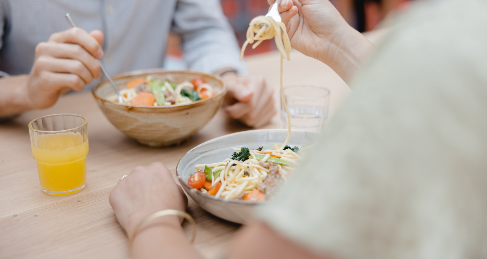 Zwei Personen essen Pasta aus Schüsseln, daneben sind zwei Gläser mit Orangensaft und Wasser