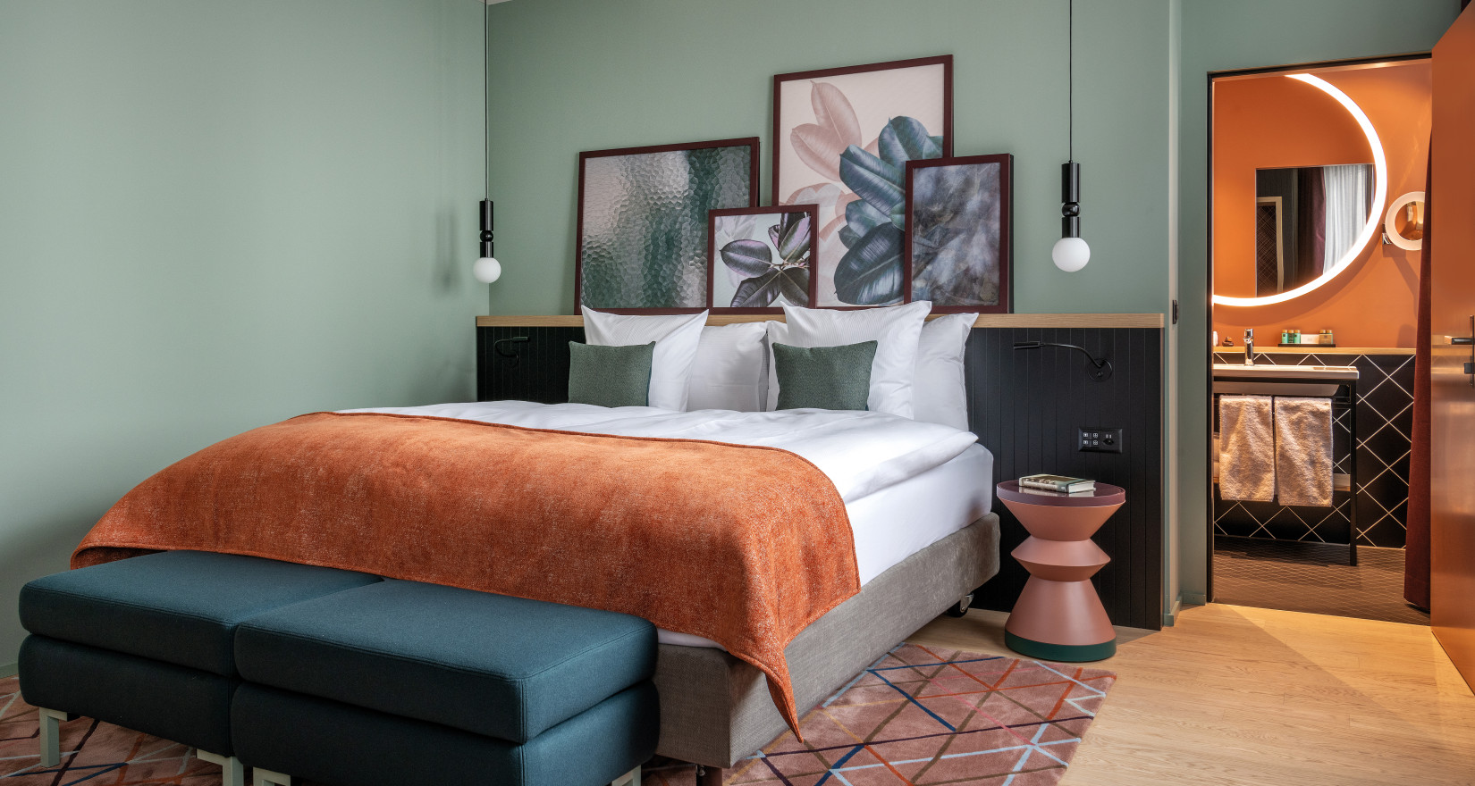 Hotelzimmer mit grüner Rückwand und einem Doppelbett mit orangem Überwurf, passend dazu ein Teppich