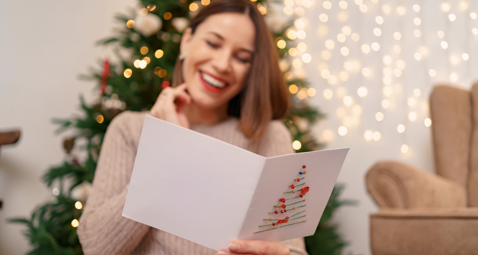 Frau liest Weihnachtskarte und lächelt dabei, hinter ihr ein Weihnachtsbaum und Lichterglanz