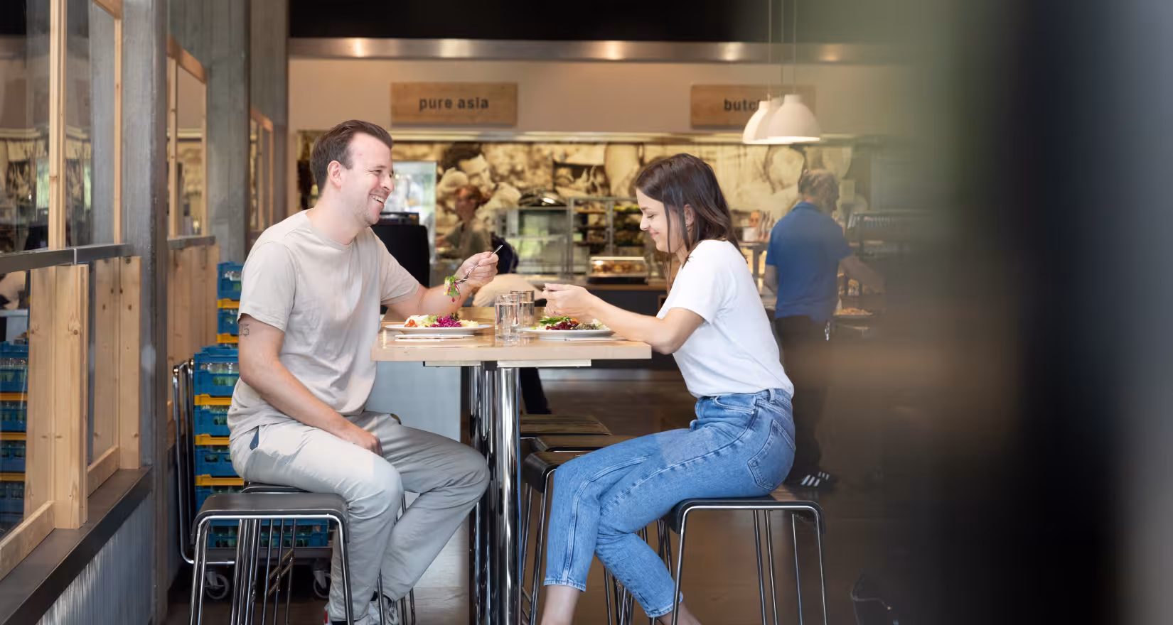 Zwei Personen sitzen an einem Hochtisch beim Mittagessen, hinter Ihnen Uniatmosphäre