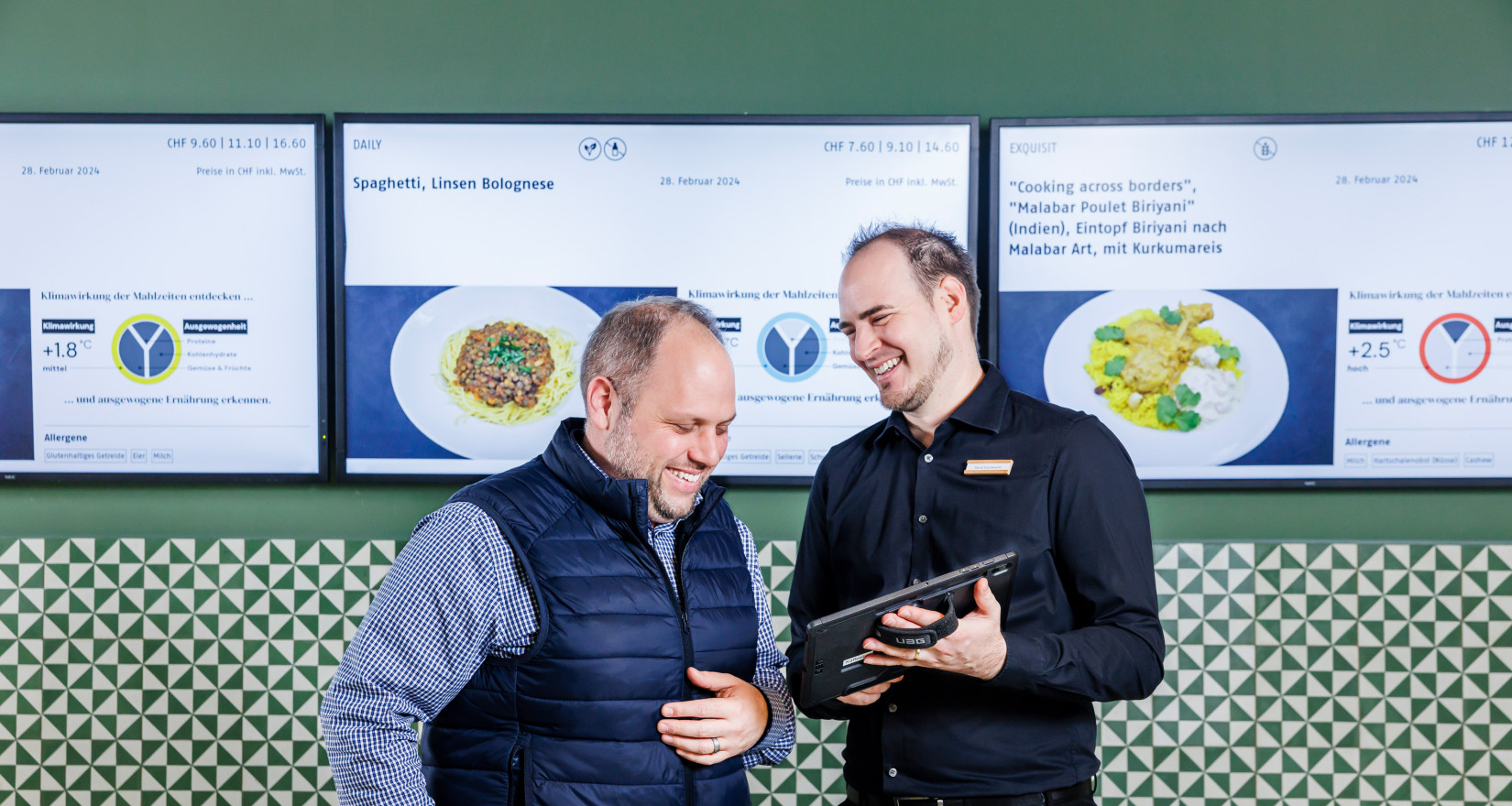 Zwei Männer stehen vor digitalen Screens und besprechen etwas auf einem Tabler