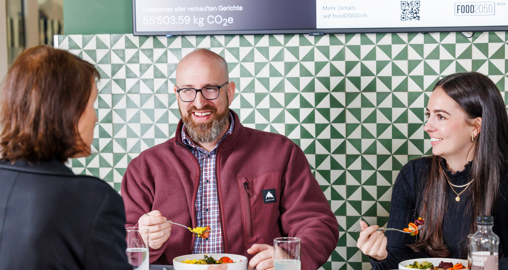 Drei Personen essen zu Mittag und sitzen vor einem Screen mit Infos zur Klimawirkung