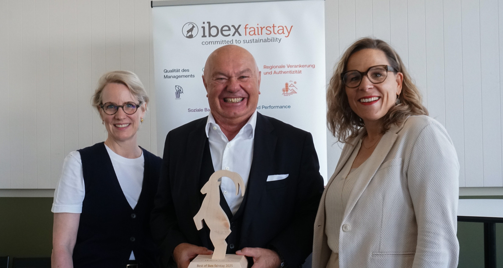 Drei Personen lächeln in die Kamera, der Mann in der Mitte hält die Auszeichnung von ibex fairstay