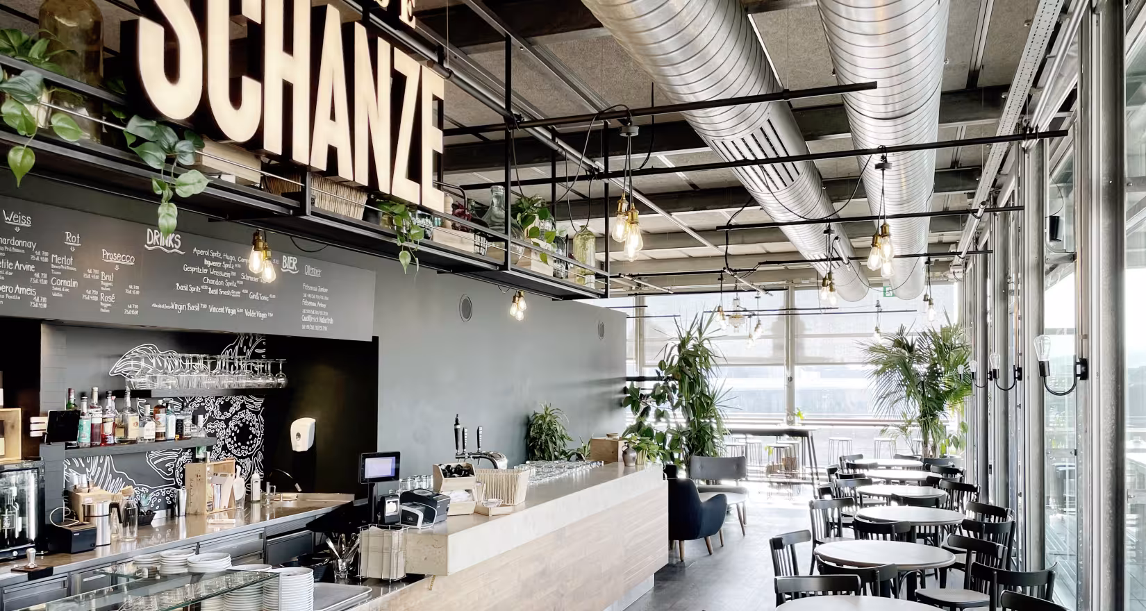 Ansicht Café-Bar mit grossem Logo Grosse Schanze, kleinen runden Tischen und Stühlen