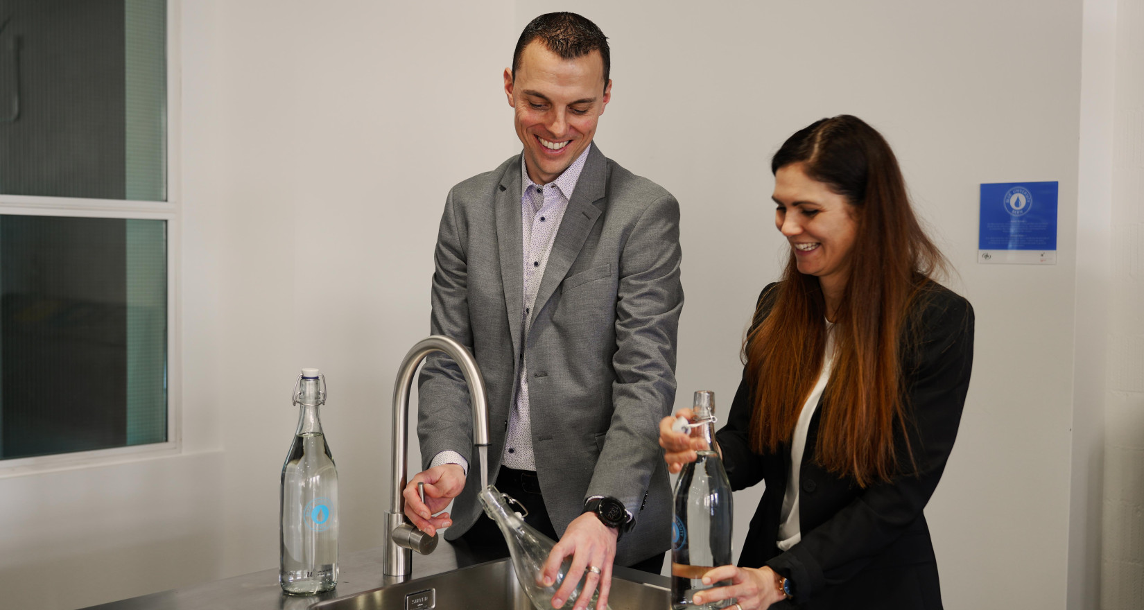 Zwei Personen bedienen sich lachend an einem Trinkbrunnen in einer Mensa