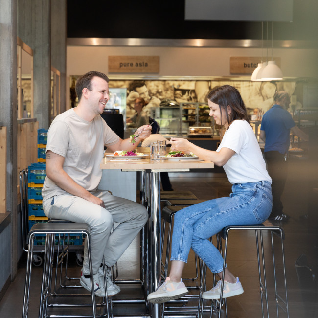 Zwei Personen sitzen an einem Hochtisch beim Mittagessen, hinter Ihnen Uniatmosphäre