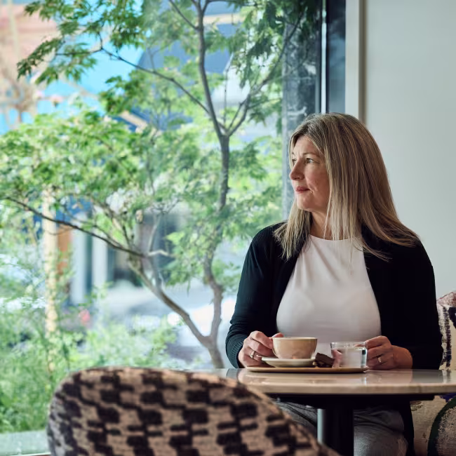 Frau sitzt in einem Kaffee und blickt nach draussen, hinter ihr der Blick ins Grüne