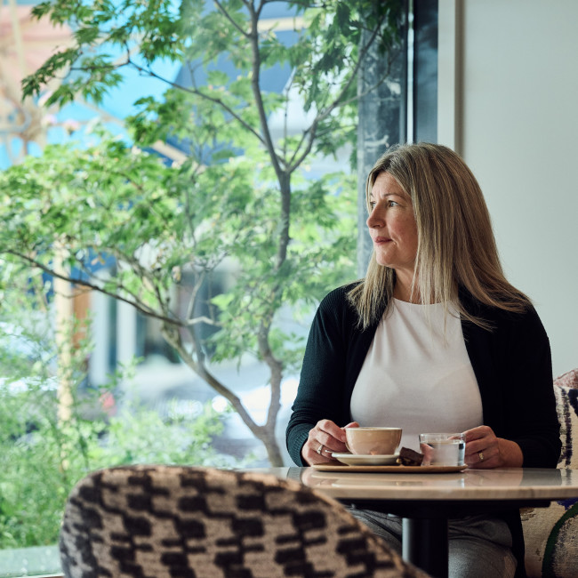 Frau sitzt in einem Kaffee und blickt nach draussen, hinter ihr der Blick ins Grüne