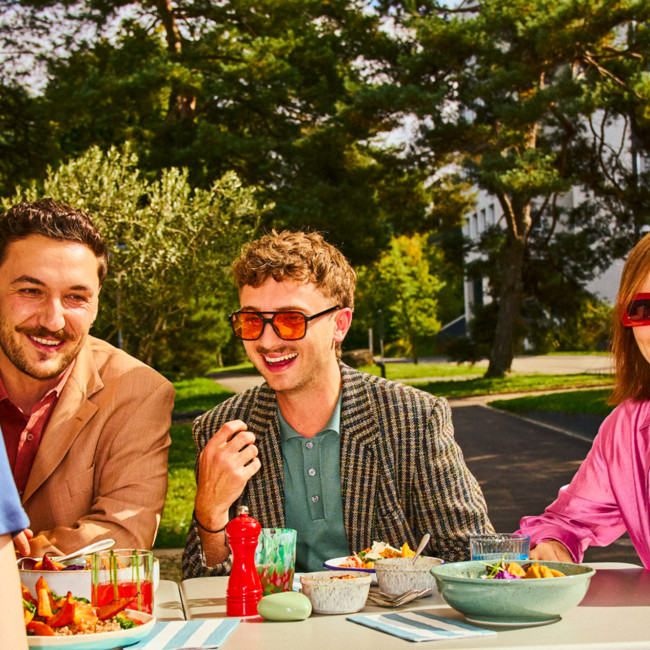 Vier Personen sitzen an einem gedeckten Tisch im Freien, essen gemeinsam und sind von viel Grün und Bäumen umgeben; auf dem Tisch stehen bunte Salate, Getränke und Geschirr in sommerlicher Atmosphäre.