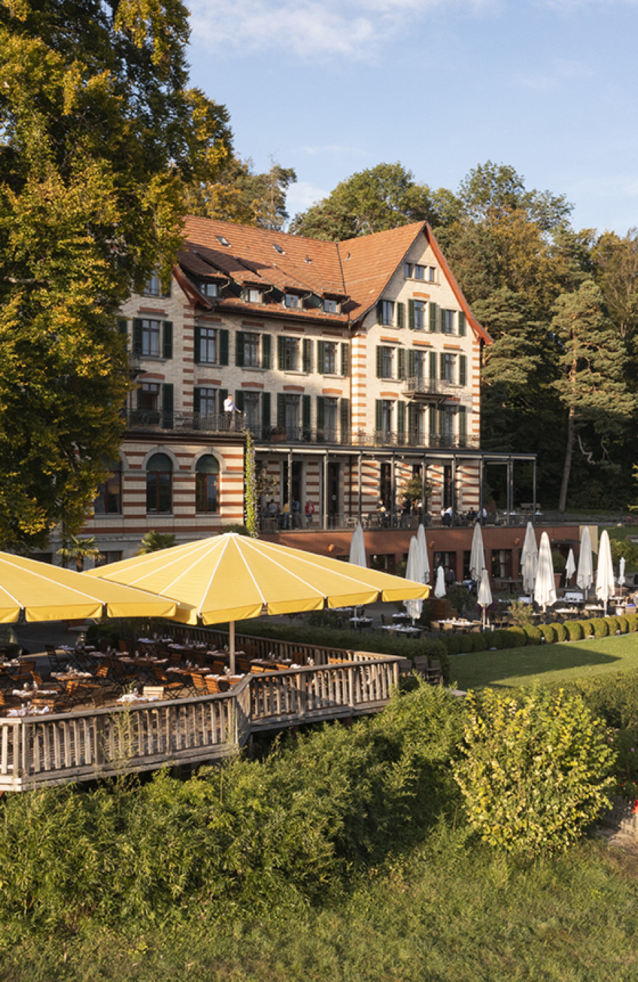 Hotel Aussenansicht mit Sonne, Grünfläche darum herum und Holzterrasse mit gelben Sonnenschirmen