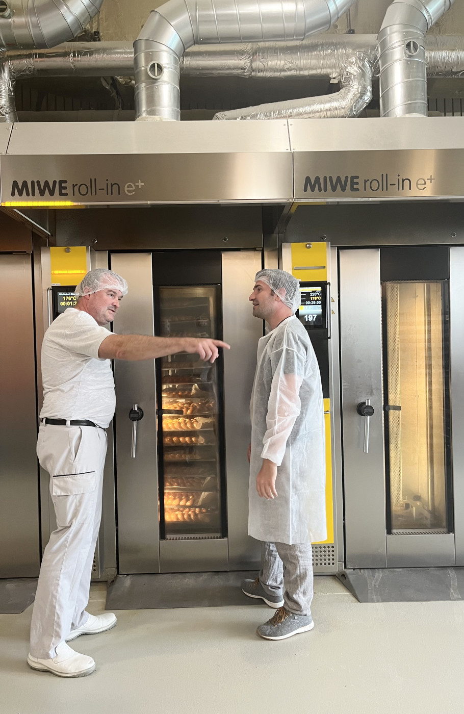 Zwei Männer in Bäckereiuniform vor grossen Industrieöfen