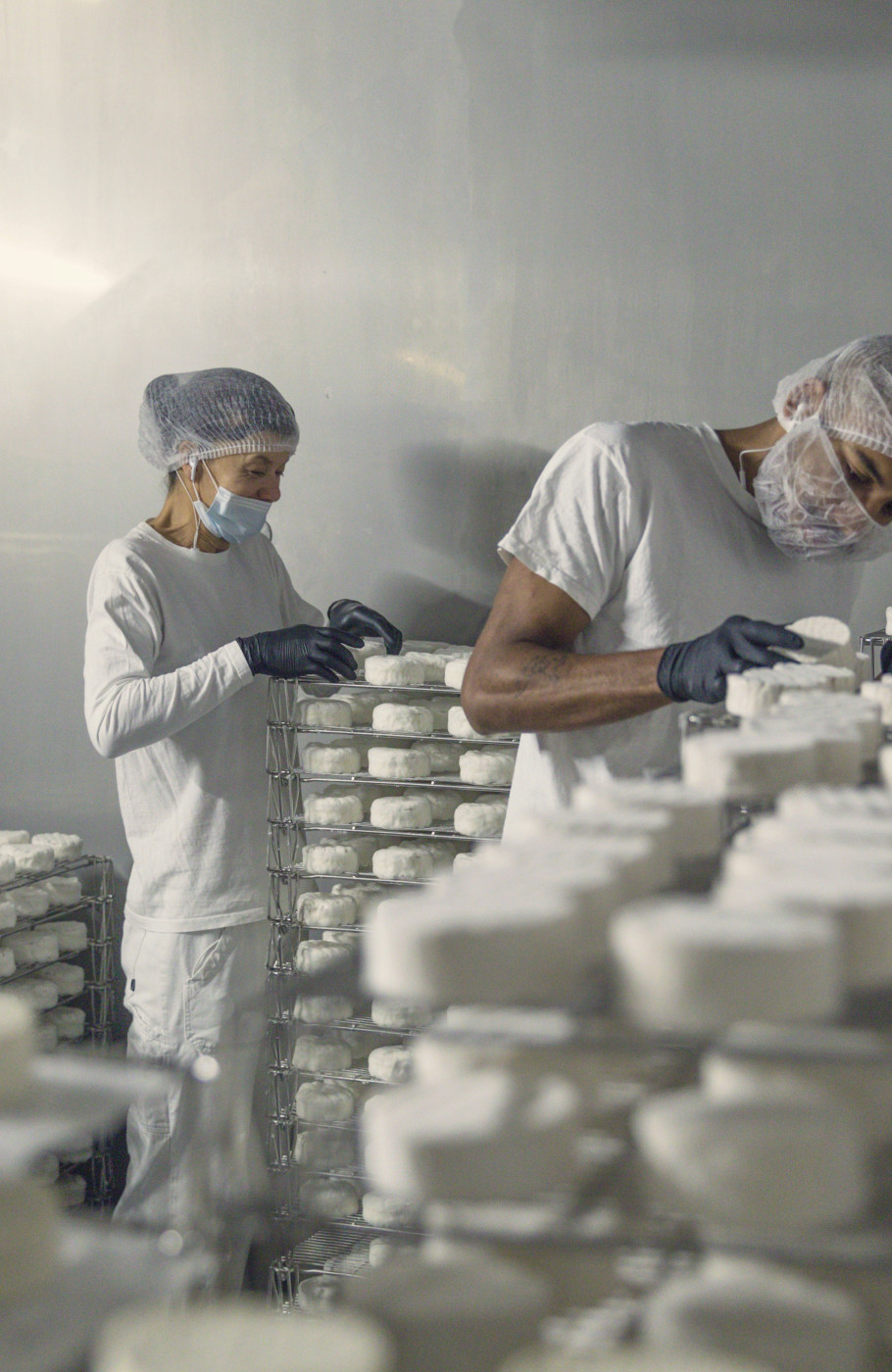 Ein Mann und eine Frau in Hygieneanzügen mit Gesichtsmasken und Hauben, arbeiten in einem Kühlraum mit Regalen voller weissem Käse