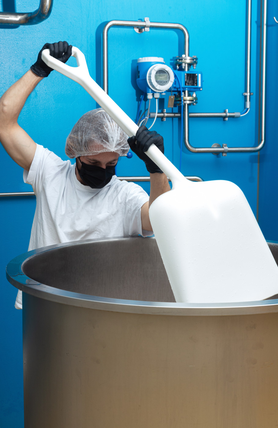 Männ in Hygieneanzug mit Maske und Haube in einer Produktion rührt mit einer grossen Schaufel in einem Metallzuber
