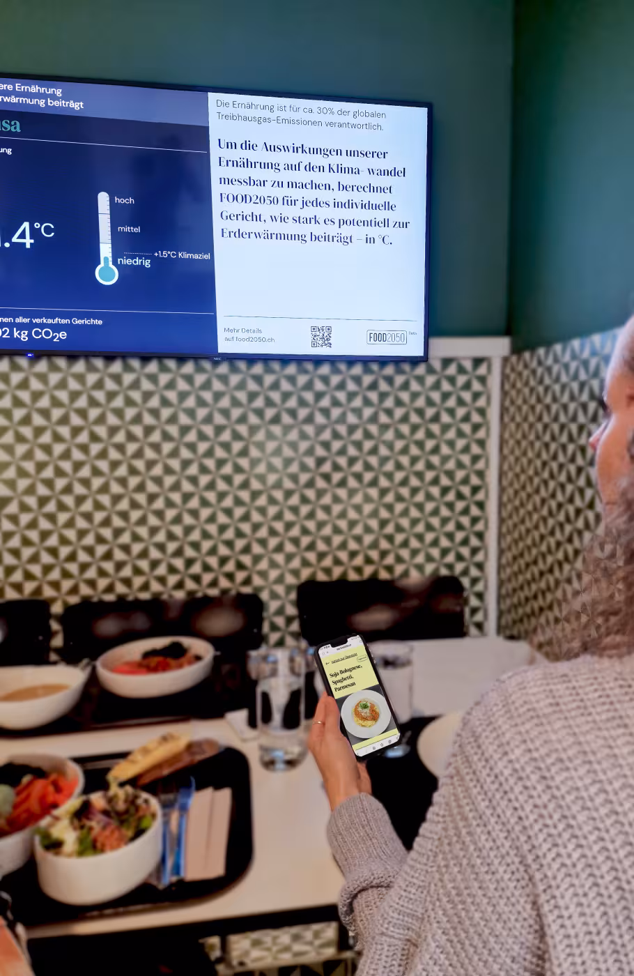 Frau sitzend vor einem Tisch mit Speisen, Bowl und Spaghetti, sie schaut auf einen Screen mit Informationen zur Klimawirkung der Menüs