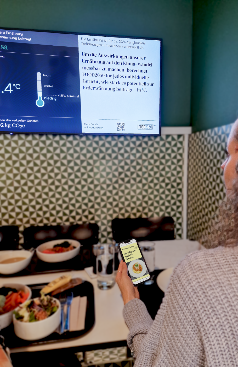 Frau sitzend vor einem Tisch mit Speisen, Bowl und Spaghetti, sie schaut auf einen Screen mit Informationen zur Klimawirkung der Menüs