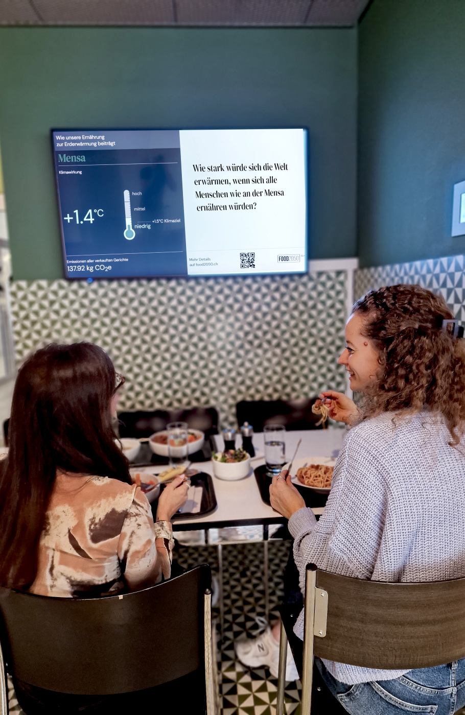 Screen an der Wand mit Informationen zum Erderwärmungspotenzial der Menüs, davor ein Tisch mit zwei Frauen beim Essen