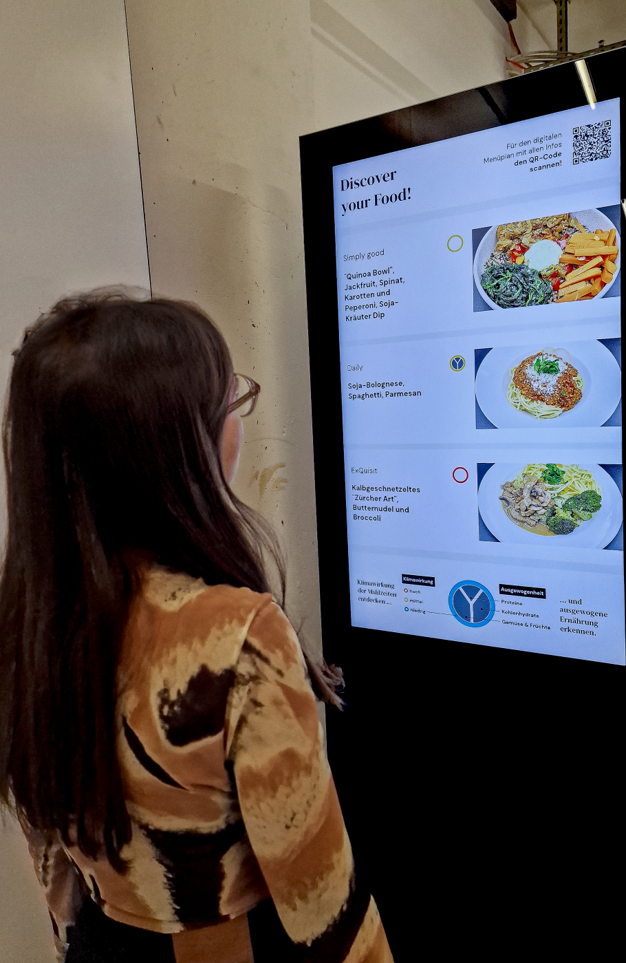 Digitale Menükarte auf einem Screen zeigt Tagesangebot an, junge Frau mit braunem Haar steht davor