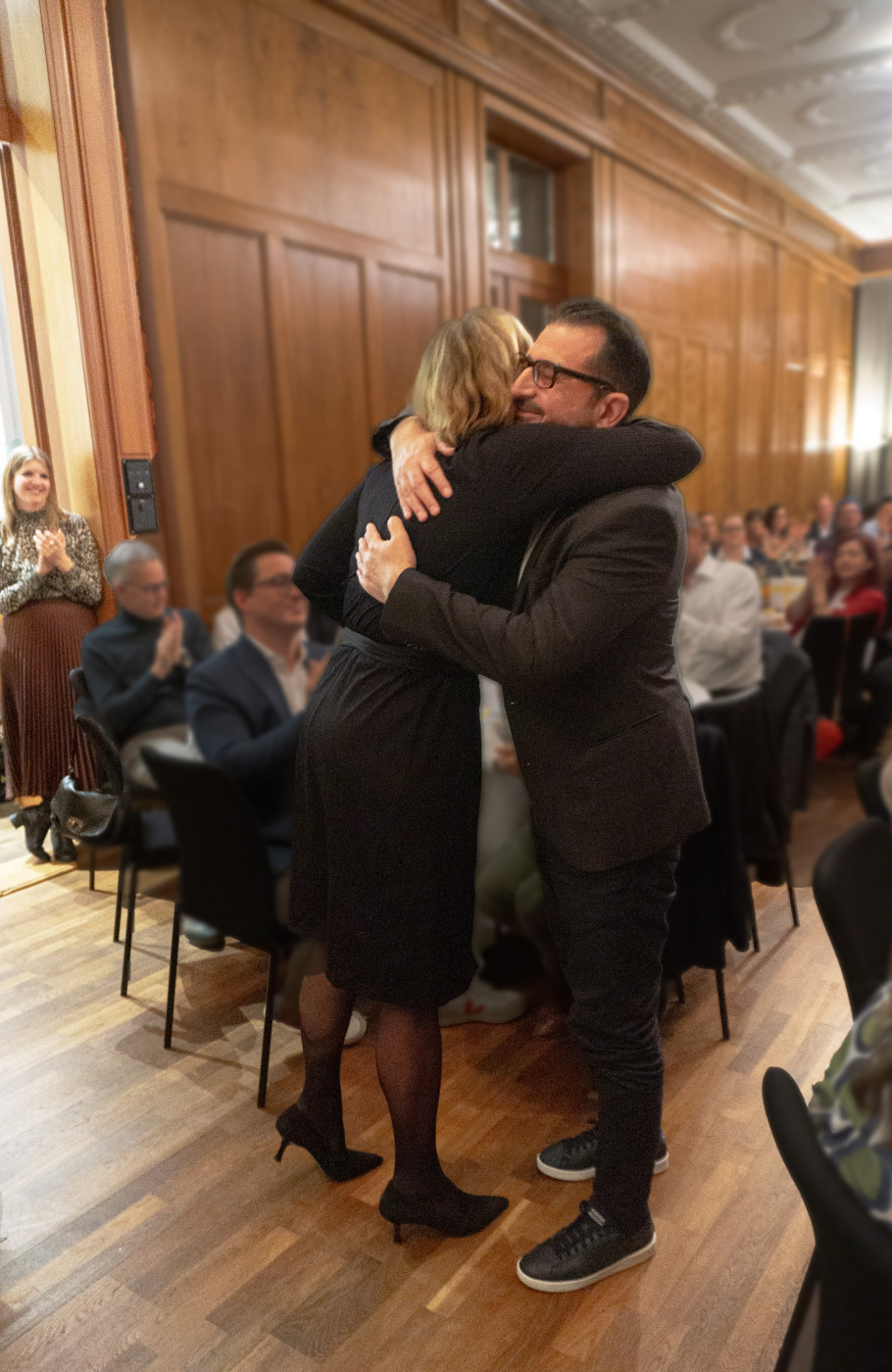 Zwei Personen umarmen sich, im Hintergrund sitzen Menschen an grossen, runden weissgedeckten Tischen