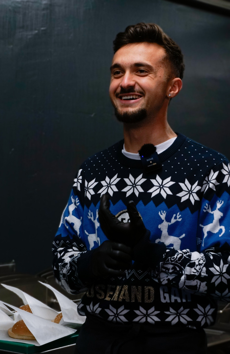 Junger Mann mit einem Weihnachtspulli zieht sich lachend Kochhandschuhe an