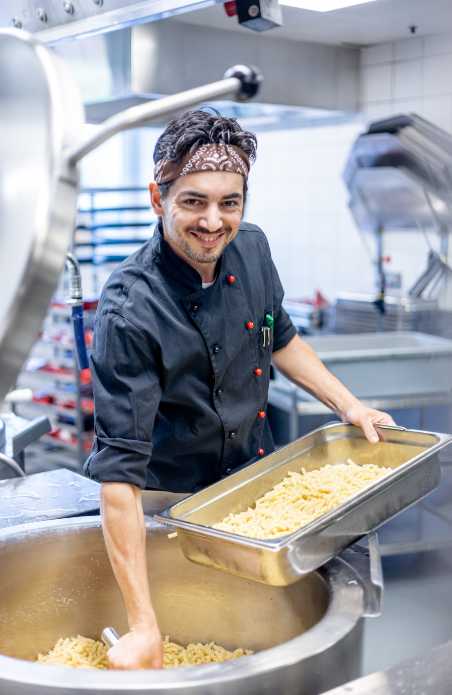 Junger Mann in schwarzer Kochuniform schöpft Pasta aus einem Gastrotopf