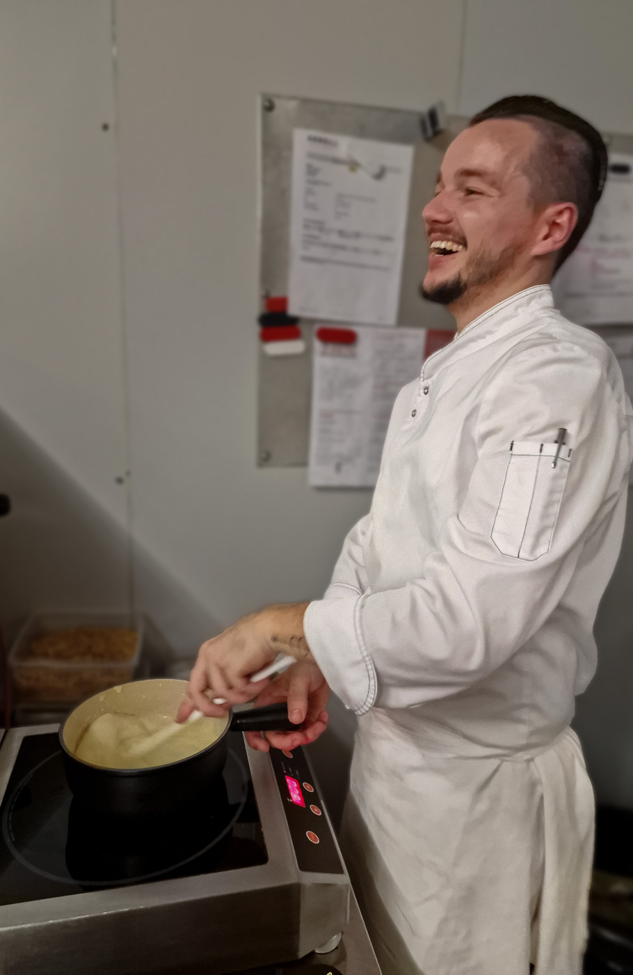 Männlicher Koch rührt lachend in einer Pfanne Fondue auf einem Herd an 