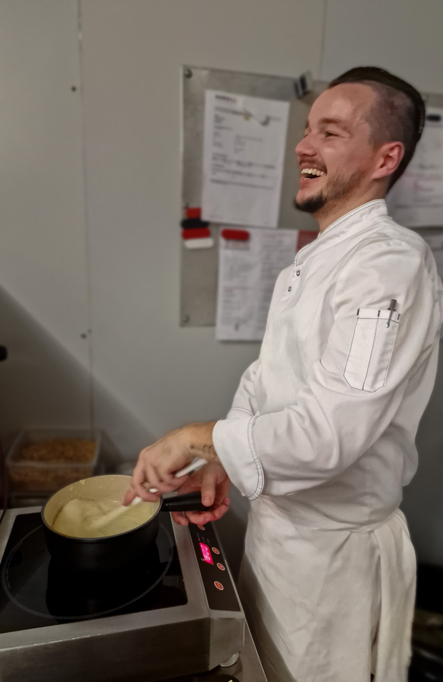 Männlicher Koch rührt lachend in einer Pfanne Fondue auf einem Herd an