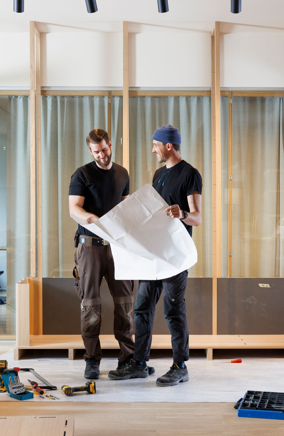 Zwei Männer prüfen einen Bauplan vor einem Holzregal