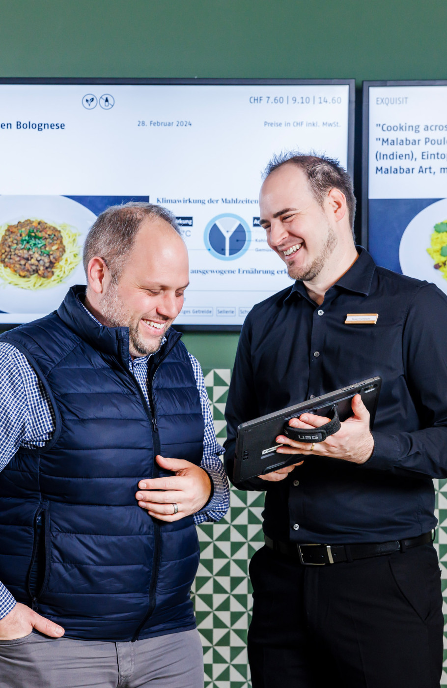Zwei Männer stehen vor digitalen Screens und besprechen etwas auf einem Tabler