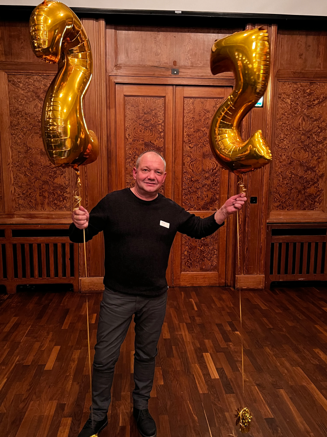 Mann blickt stehend in die Kamera und hält goldene Ballons mit einer 25