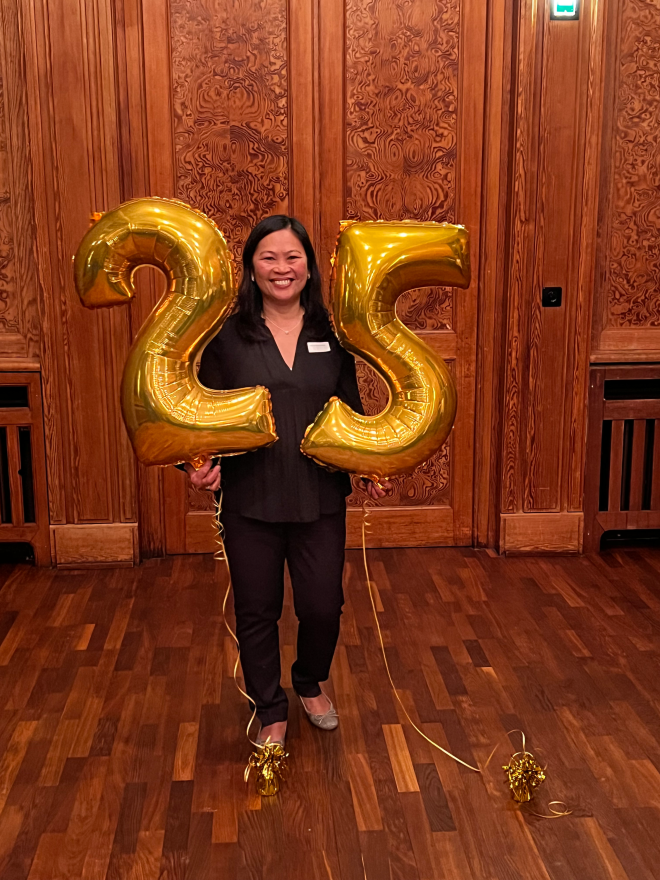 Frau blickt stehend in die Kamera und hält goldene Ballons mit einer 25