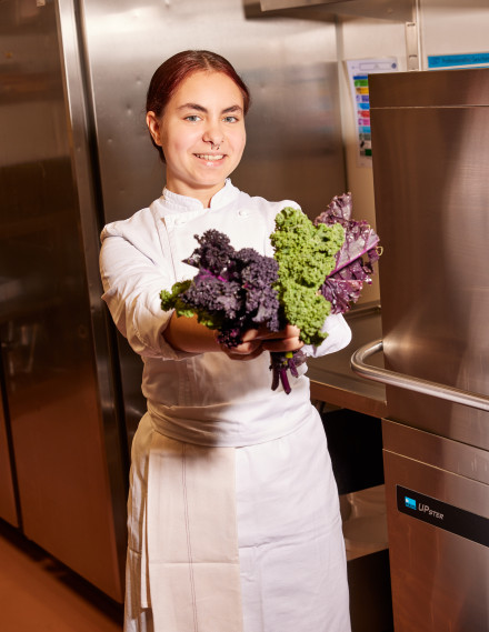 Junge Frau steht in Küchenkleidung in einer Gastronomieküche und hält Blätter