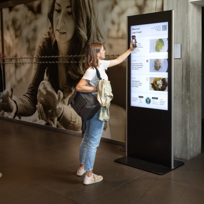 junge Frau scannt mit ihrem Handy einen digitalen Screen mit einem Foodangebot