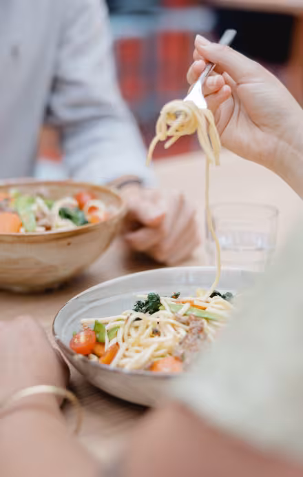 Zwei Personen essen Pasta aus Schüsseln, daneben sind zwei Gläser mit Orangensaft und Wasser