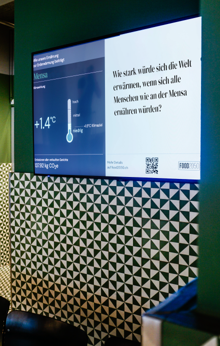 Live Ticker auf einem Screen zeigt Umweltauswirkungen der Menüs vor Ort auf, hängt an einer grünen Wand