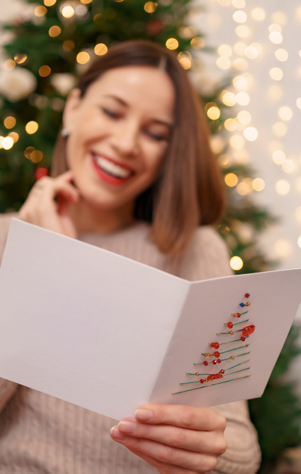 Frau liest Weihnachtskarte und lächelt dabei, hinter ihr ein Weihnachtsbaum und Lichterglanz