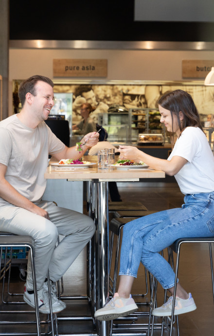 Zwei Personen sitzen an einem Hochtisch beim Mittagessen, hinter Ihnen Uniatmosphäre