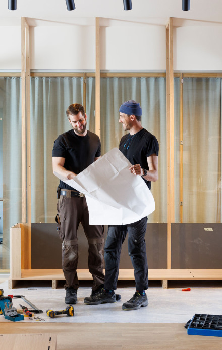 Zwei Männer prüfen einen Bauplan vor einem Holzregal