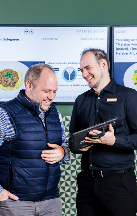 Zwei Männer stehen vor digitalen Screens und besprechen etwas auf einem Tabler