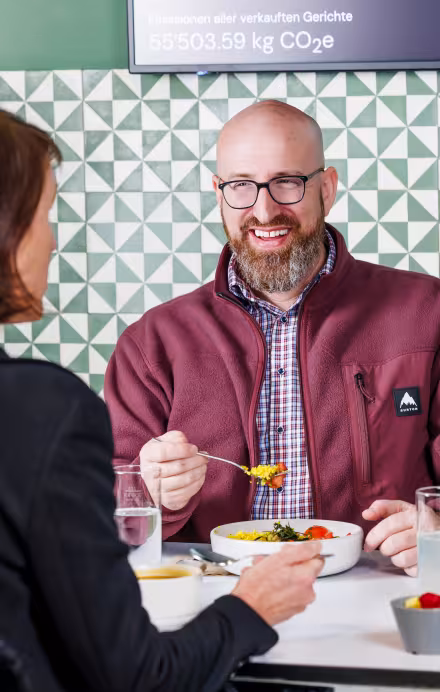 Drei Personen essen zu Mittag und sitzen vor einem Screen mit Infos zur Klimawirkung