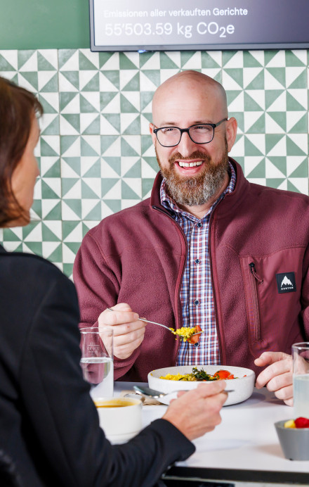Drei Personen essen zu Mittag und sitzen vor einem Screen mit Infos zur Klimawirkung