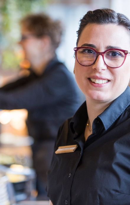 Frau lächelt in die Kamera, im Hintergrund ist ein Restaurant zu sehen